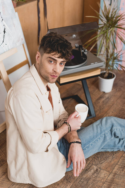 handsome artist sitting on floor with coffee cup and looking at camera - Foto, immagini
