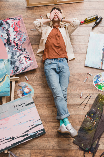 vista aérea do artista cansado deitado no chão de madeira no estúdio de pintura e de mãos dadas no rosto
 - Foto, Imagem