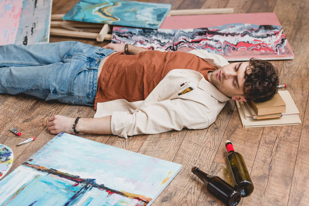 exhausted artist lying on wooden floor, surrounded with paintings, draw utensils and empty bottles - Photo, Image