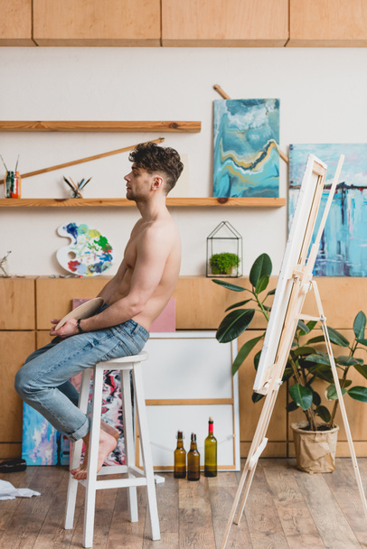 guapo artista semidesnudo en vaqueros azules sentado en la silla alta en el estudio de pintura
 - Foto, Imagen