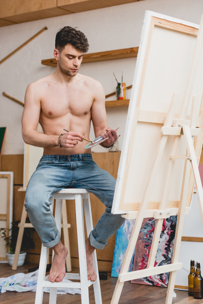 handsome half-naked artist mixing colors on palette while sitting on high chair in gallery - Photo, Image