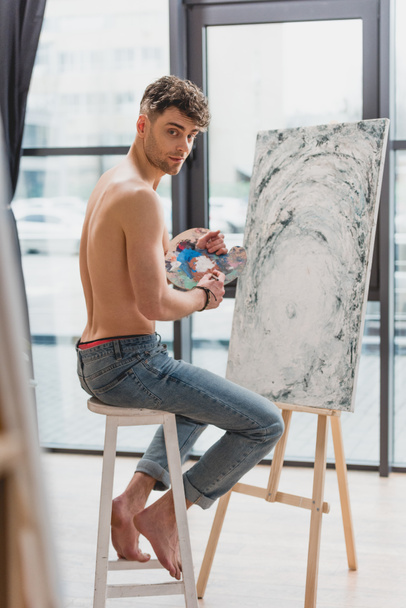 selective focus of good-looking half-naked artist looking at camera while sitting on high chair near easel with painting - Foto, afbeelding