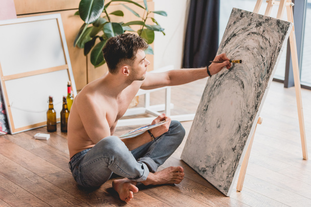 handsome half-naked artist sitting with crossed legs on floor and painting picture on canvas - Photo, Image