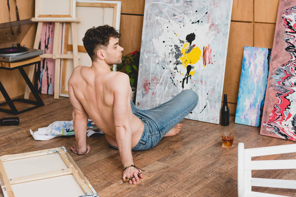 красивий напівголий художник сидить на підлозі перед полотном з малюнком
 - Фото, зображення