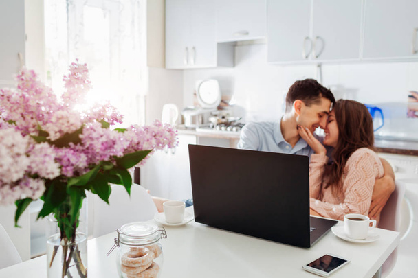 Happy young couple using laptop while having breakfast in modern kitchen. Young man and woman drink coffee and hug - Photo, Image