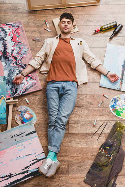 tired artist lying of floor, surrounded with paintings and draw utensils, and looking at camera - Photo, Image