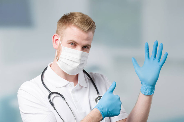 doctor with medical face mask and stethoscope puts on his medical gloves and shows thumb up - Foto, immagini