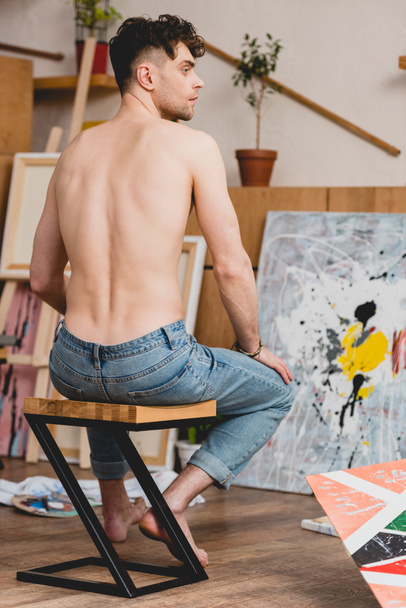 back view of half-naked artist in blue jeans sitting on chair in painting studio - Photo, Image