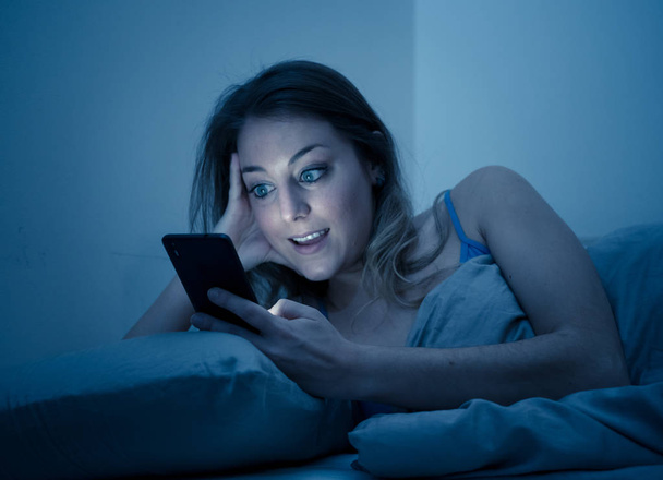 Happy young beautiful girl talking and surfing on the internet using her smart phone. Cheerful and joyful female video chatting late at night in relax and casual mood. In mobile addiction concept. - Fotoğraf, Görsel