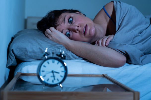 Sleepless and desperate beautiful caucasian woman awake at night not able to sleep, feeling frustrated and worried looking at clock suffering from insomnia in sleep disorder concept. - Photo, Image