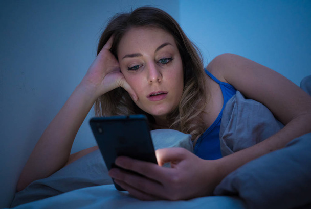 Addicted young beautiful woman in bed, chatting and surfing on the internet using her smart phone sleepy, dull and tired late at night in mobile addiction and technology overuse concept. - Фото, зображення