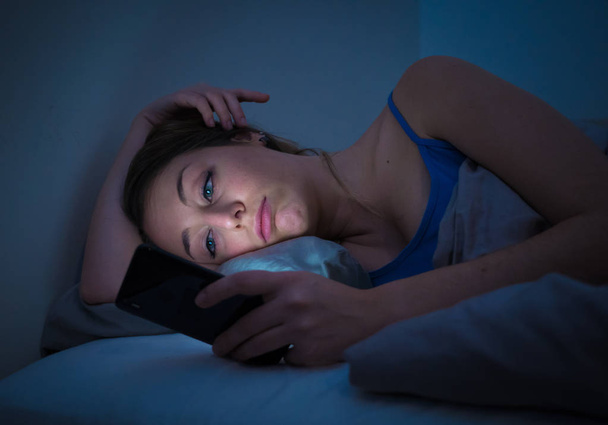 Addicted young beautiful woman in bed, chatting and surfing on the internet using her smart phone sleepy, dull and tired late at night in mobile addiction and technology overuse concept. - Foto, imagen