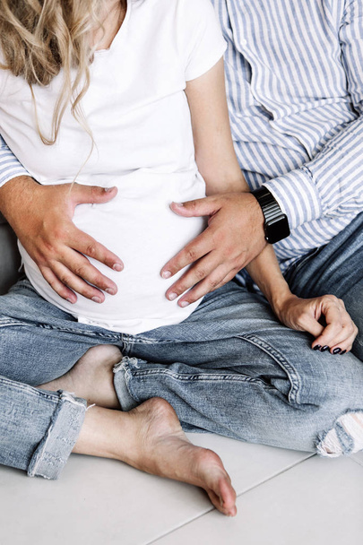 Pregnancy, a couple in light jeans and white T-shirts are sitting on the floor and the man is holding a pregnant belly - Photo, Image