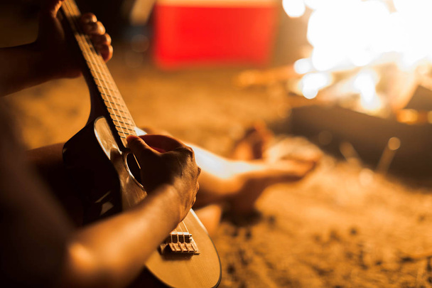 Una mujer músico tocando la guitarra ukelele junto a una fogata en la playa
. - Foto, Imagen