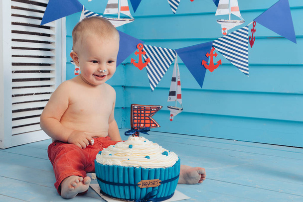 decoration for the boys birthday, smash the cake in a nautical marine style. stylized birthday ship photo shoot. Cheerful boy eats and break a cake with his hands on the first holiday - Photo, Image
