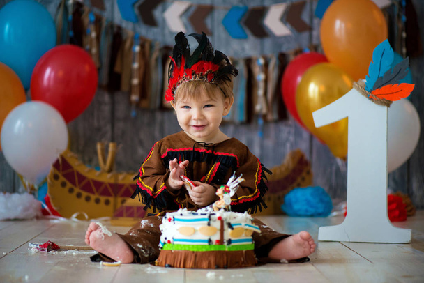 little cute boy in costume of American Indian Apache Chief sits and smash cake. Children's holiday, happy Birthday 1 year, photo zone decoration - Fotoğraf, Görsel