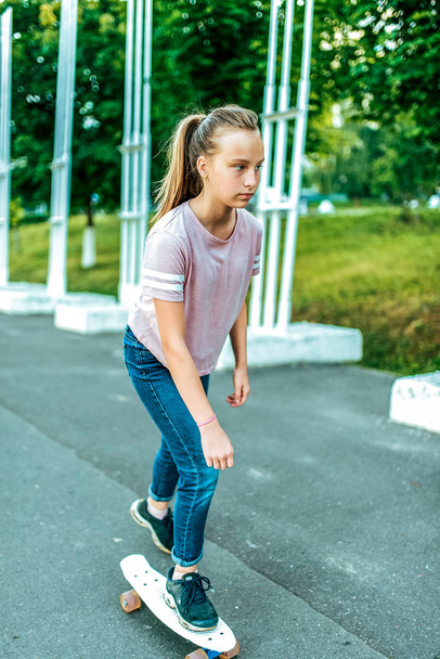 A girl is a schoolgirl of 12-15 years old, riding a skateboard in the summer in the city, an asphalt road. Casual clothes, jeans sneakers T-shirt. - Фото, зображення