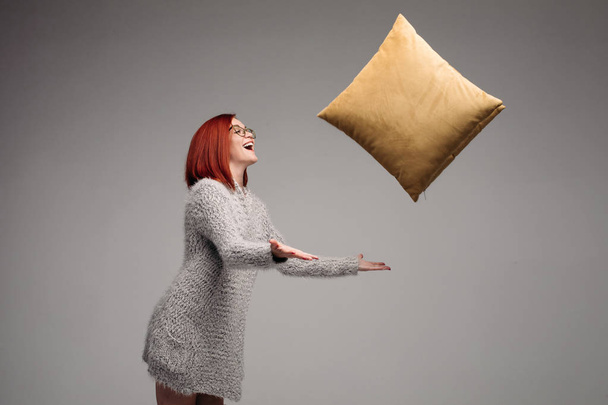 Red haired woman in gray sweater holding and giving at side yellow pillow. - Photo, Image