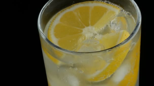 Sliced lemon in a long glass with ice cubes and cold soda on a black background close up view. Refreshing Lemonade Bubbles. - Footage, Video