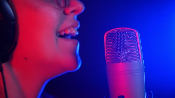 Μια νεαρή χαμογελαστή γυναίκα με ακουστικά που τραγουδούν στο στούντιο. Φωτισμός νέον - Πλάνα, βίντεο