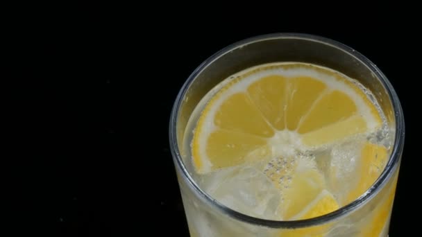 Gesneden citroen in een lang glas met ijsblokjes en koude soda op een zwarte achtergrond close-up te bekijken. Verfrissende limonade bubbels. - Video