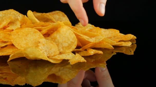 Womens hands take potato chips lie on a mirror surface randomly scattered on a black background. Harmful food, fast food. - Footage, Video