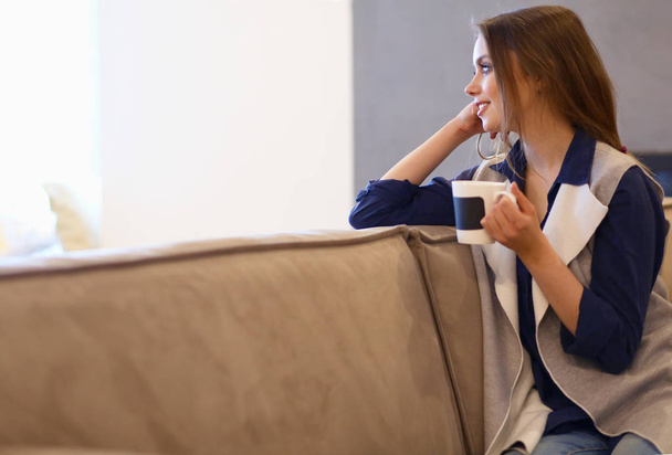 Young woman sitting on comfortable sofa with cup on coffee in hands in white room - Foto, imagen