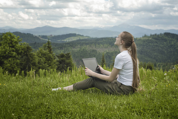 Beautiful dreaming girl with a laptop sitting on green grass on a background of mountains - Photo, Image