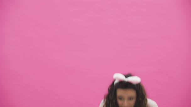 Beautiful young girl standing on a pink background. During this, there are ears of rabbits on the head. - Video