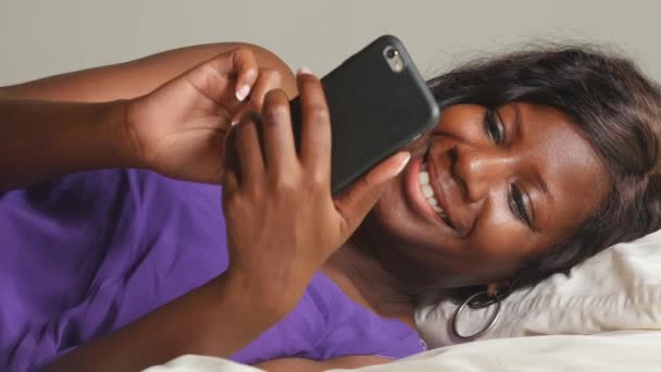 Mladá krásná a šťastná 30s černá africká americká žena ležící uvolněně na posteli pomocí internetu mobilní telefon s úsměvem veselá sítě sociální média obchodní nebo Pomaturitní studium doma ložnice - Záběry, video