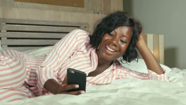 молодая красивая и счастливая чернокожая афроамериканка в пижаме лежала расслабленной и уютной на кровати, общаясь с интернет-телефоном онлайн знакомства или наслаждаясь социальными сетями улыбаясь весело
 - Кадры, видео