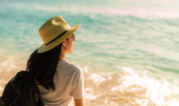 Вид сзади счастливой молодой азиатской женщины в повседневном стиле с соломенной шляпой и рюкзаком. Отдохните и насладитесь отдыхом на тропическом райском пляже. Летние вибрации. Отдых и наслаждение на тропическом пляже
. - Фото, изображение