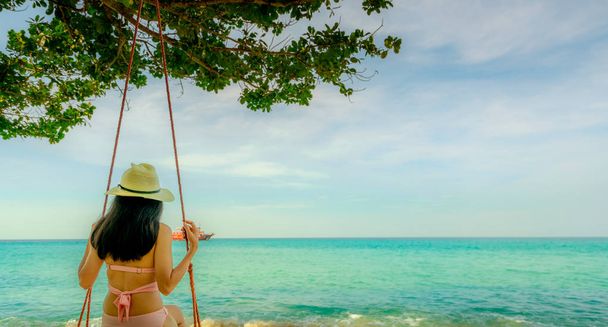 Ασιατικές γυναίκα φορούν μαγιό και καπέλο swing την ταλάντευση σε παραλία με άμμο και αναζητούν όμορφο τροπικό παράδεισο θάλασσας και ουρανού ηλιόλουστη ημέρα. Καλοκαιρινές διακοπές. Vibes καλοκαίρι. Κορίτσι απολαμβάνοντας και χαλαρώνοντας στις διακοπές - Φωτογραφία, εικόνα
