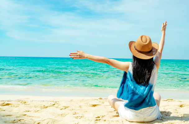 Happy young woman wear straw hat sit and raised hand at sand beach. Relaxing and enjoy holiday at tropical paradise beach with green water. Girl in summer vacation. Summer vibes. Carefree concept. - Photo, Image