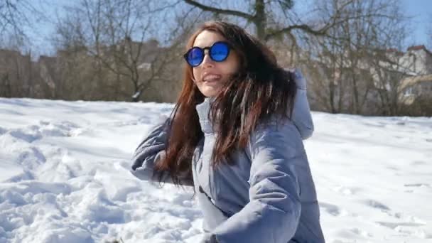 Niesamowita dziewczyna dorosłych w niebieskie lustrzane okulary i proste ciemne włosy rzucanie śnieżkami grając z kimś na zewnątrz w parku w godzinach porannych w słoneczne, ale zimny dzień. Koncepcja gier zimowych - Materiał filmowy, wideo