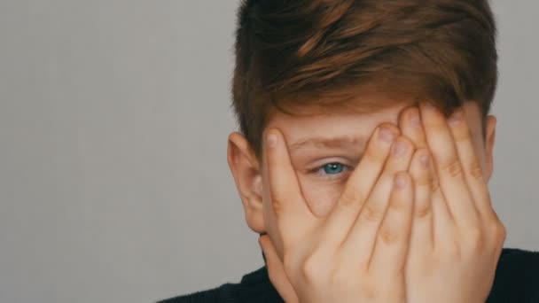 Portrait of funny and scared redhead and blue-eyed teenager boy looks around and into camera through the fingers of his hands on his face on a white background. Emotions of teenagers - Footage, Video