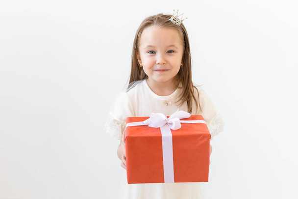 Концепция праздника и подарки - Маленькая девочка улыбается и держит красный подарочный коробок
 - Фото, изображение