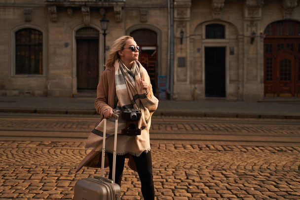 Young beautiful blond woman tourist with a wheeled travel bag and vintage film camera standing on Rynok square in Lviv, Ukraine in the morning - Fotografie, Obrázek
