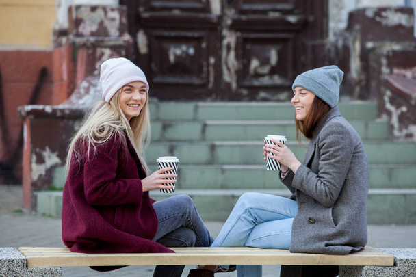 Two sisters relaxing and drinking coffee. Coffee break. Coffee to go.Outdoors fashion portrait of two young beautiful girls drinking coffee. Coffee to go.Two friends relaxing and drinking coffee .Coffee break.Coffee to go - Photo, Image