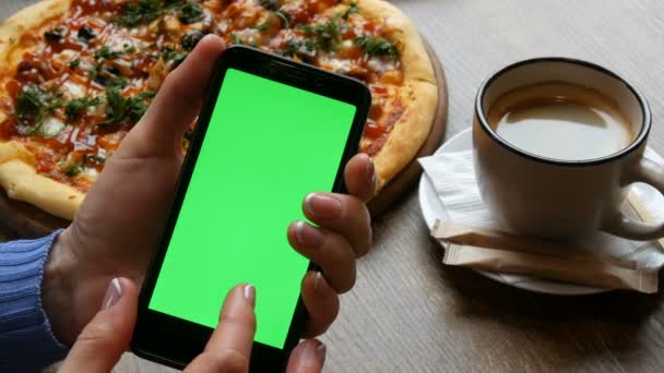 Tecla de croma o pantalla verde en un teléfono inteligente negro en manos femeninas con una manicura bien arreglada en el fondo de una pizza grande y una taza de café
 - Metraje, vídeo