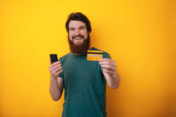 Photo of emotional excited young man standing isolated over yellow wall background holding credit card using mobile phone. Looking camera. - Photo, Image