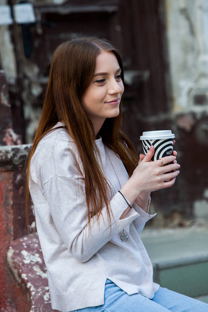Kaunis nainen tilalla paperi kahvikuppi kaupungissaNuori tyylikäs nainen juo kahvia mennä kaupungin kadulla
  - Valokuva, kuva