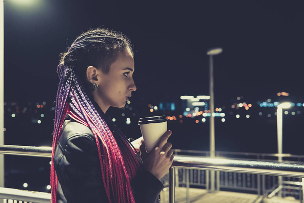 gyönyörű divatos fiatal nő, kávé, az éjszakai város. egy fiatal nő iszik kávét keresztül szalmát, egy eldobható műanyag pohár - Fotó, kép