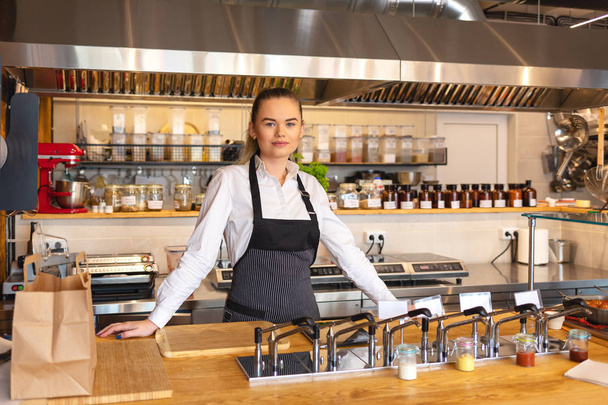 Ritratto di giovane donna in piedi dietro il bancone della cucina in piccolo ristorante, proprietario del ristorante che indossa un grembiule guardando fiducioso alla fotocamera sorridente
 - Foto, immagini