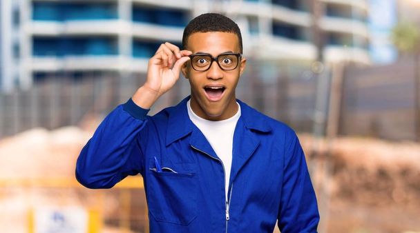 Young afro american worker man with glasses and surprised in a construction site - Foto, afbeelding