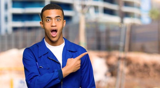 Young afro american worker man surprised and pointing side in a construction site - Photo, Image