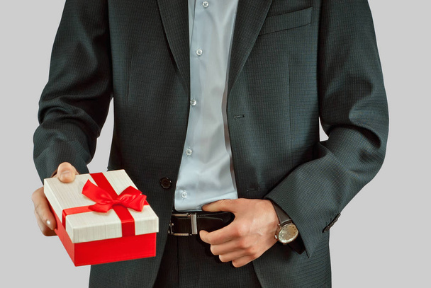 Un hombre con traje tiene una caja de regalo en sus manos. Caja con una cinta roja y un lazo
. - Foto, Imagen