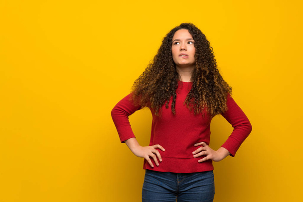 Teenager girl with red sweater over yellow wall with confuse face expression while bites lip - Fotó, kép
