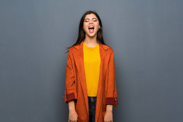 Chica adolescente con abrigo sobre pared gris gritando al frente con la boca abierta
 - Foto, imagen
