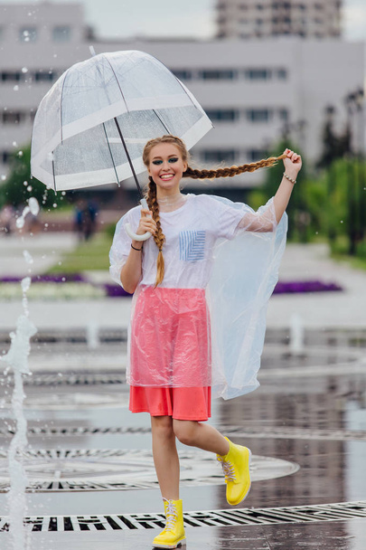 junges hübsches Mädchen mit zwei Zöpfen in gelben Stiefeln und mit transparentem Regenschirm steht in der Nähe des Brunnens. - Foto, Bild